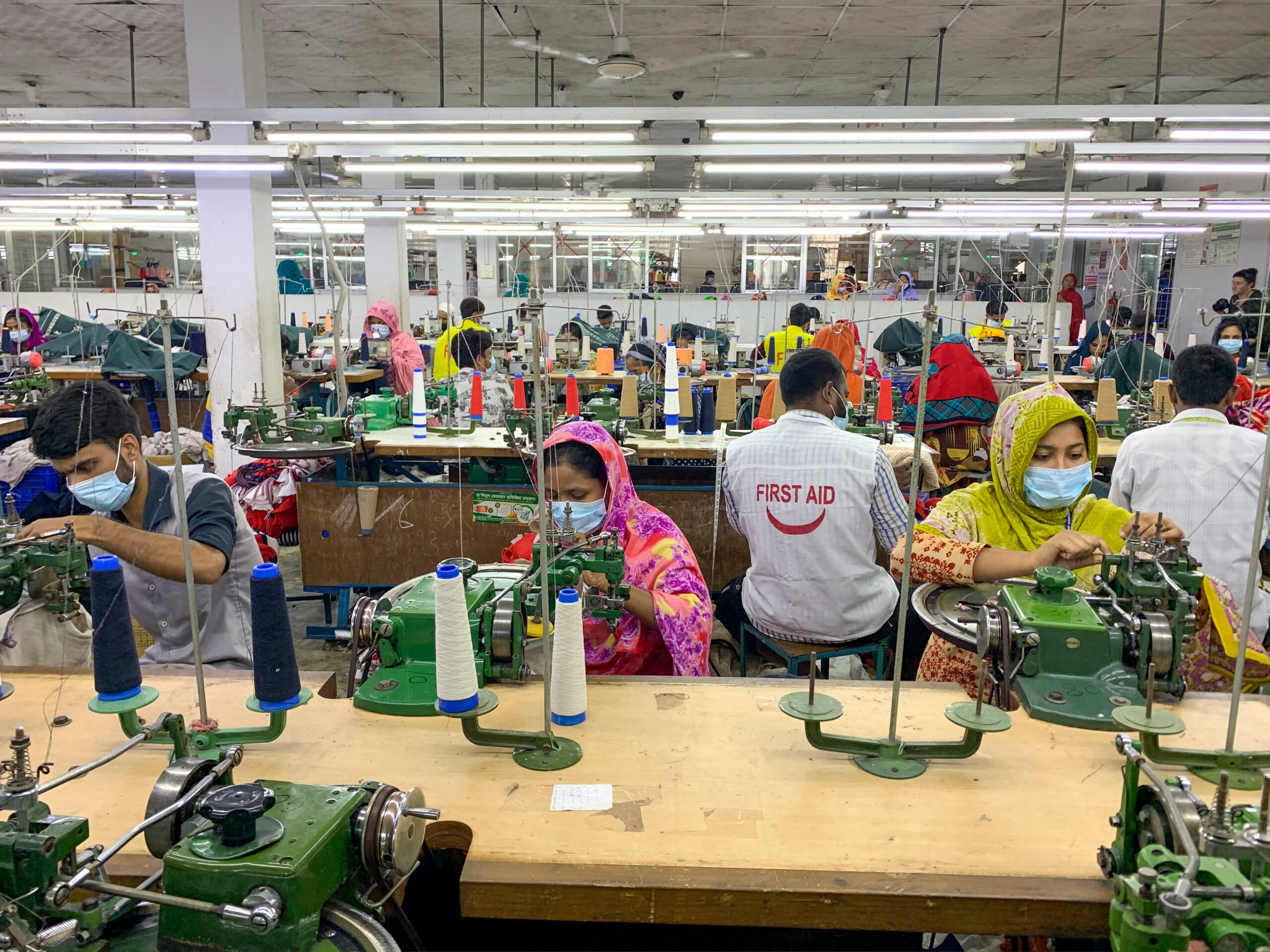 Workers operating machinery in a garment factory, two women face towards the camera though are focussed on their work. Other workers face away from the camera, one has 'First Aid' written on their bib.